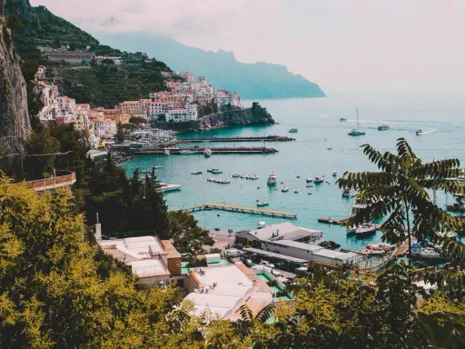 Vista panoramica di un villaggio colorato sulla Costiera Amalfitana, con barche nel porto e montagne sullo sfondo. Un invito a scoprire come visitare la Costiera Amalfitana.