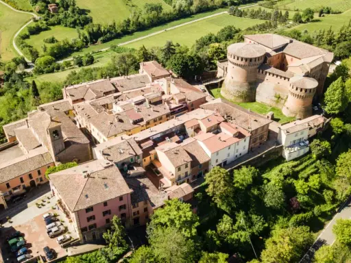 Aerial view of a medieval Italian village with a castle, terracotta roofs, and surrounding green hills.