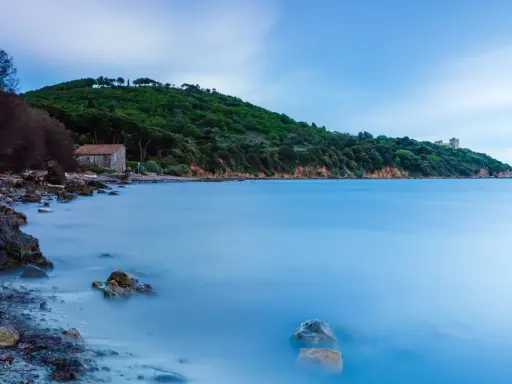 Una casa di pietra sulla spiaggia di Bengodi, con il mare calmo e una collina verdeggiante sullo sfondo.