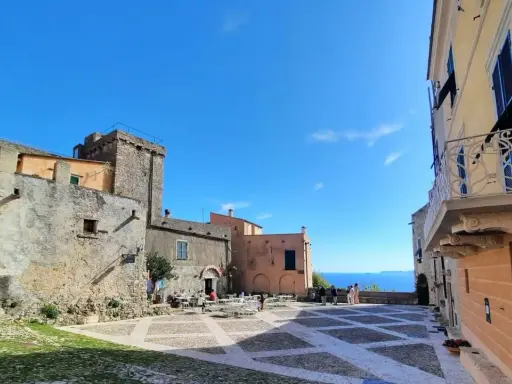 Piazza nel centro storico di Borgio Verezzi con edifici antichi, tavolini all'aperto e il mare sullo sfondo.