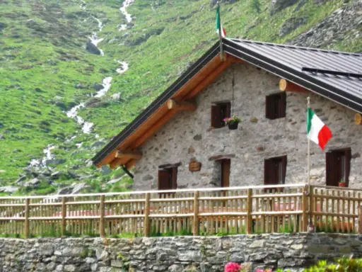 Rifugio in pietra con bandiera italiana, circondato da verdi pendii e un ruscello. Un'oasi accogliente tra i rifugi Val Camonica facili da raggiungere.