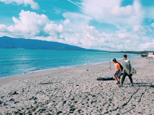 Due ragazzi passeggiano sulla spiaggia della Maremma Toscana, uno con una tavola da surf, mentre il mare azzurro lambisce la riva.