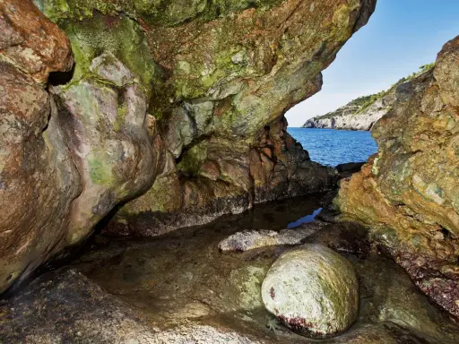 L'entrata di una grotta marina, la cala del bove, con rocce muschiose e un piccolo specchio d'acqua che riflette il blu del mare e il cielo.