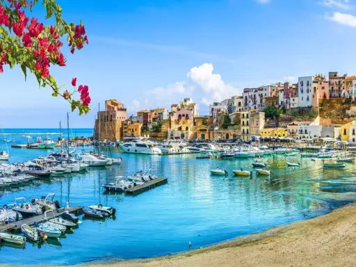 Un pittoresco porto con barche ormeggiate, case colorate su una collina e fiori rossi in primo piano, un'immagine che evoca la bellezza di Sardegna e Sicilia.