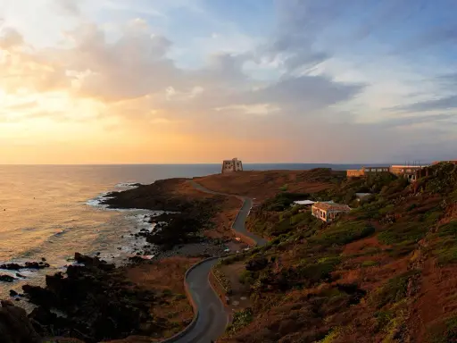 Torre di tramontana e case sparse su una collina, con mare e cielo al tramonto. Ecco cosa vedere a Ustica per un'esperienza indimenticabile.