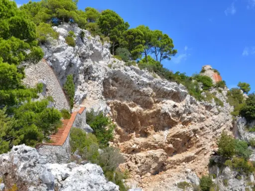 Scogliera rocciosa con una scalinata in mattoni che sale verso la cima, dove si intravede un rudere. Si immagina il **salto di Tiberio** da questo luogo suggestivo.