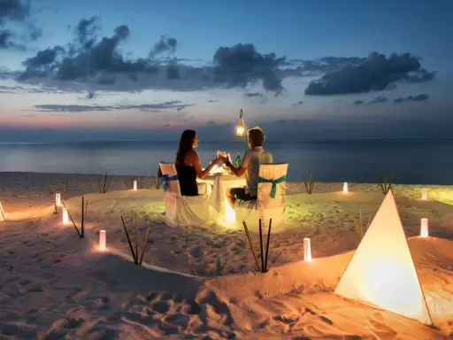 Una coppia al mare di sera, seduta a tavola per una cena romantica illuminata da lanterne e luci soffuse sulla sabbia.