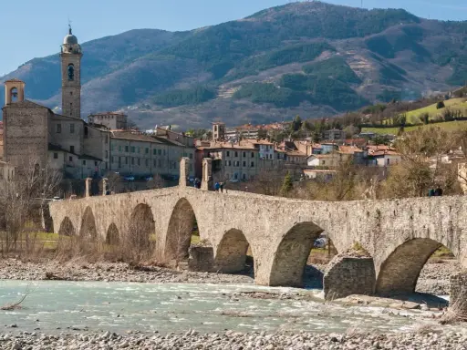 Ponte medievale in pietra attraversa un fiume, con un borgo storico e colline sullo sfondo. Un gioiello tra i borghi medievali dell'Emilia Romagna.
