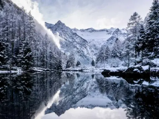 Val di Mello inverno: alberi innevati e montagne maestose si specchiano in un lago calmo, creando un paesaggio da fiaba.
