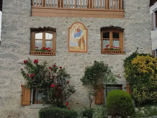 Un affresco sacro adorna un muro di pietra di un borgo trentino alto adige, con rose rosse che sbocciano rigogliose.