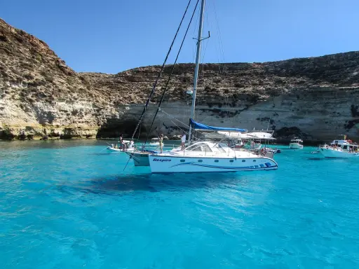 Catamarano "Respiro" in acque cristalline di un'isola sotto la Sicilia, con scogliere rocciose e altre barche.