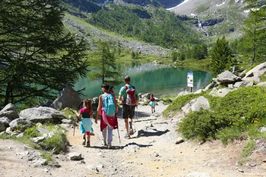 Famiglia in escursione sul sentiero di un lago alpino, con montagne verdi e un ruscello sullo sfondo. Valle d'Aosta estate.