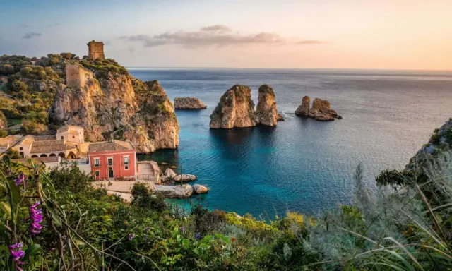 Un borgo con torri antiche e una casa rossa si affaccia su un mare bello Italia, con faraglioni che emergono dalle acque cristalline.