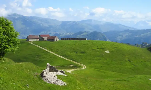 Un sentiero delle malghe serpeggia tra verdi colline, conduce a rustici edifici e si perde tra le montagne nebbiose.