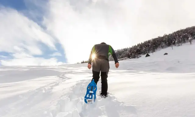 Uomo con ciaspole cammina su sentiero innevato, ideale per chi cerca dove ciaspolare in Piemonte. Sole splendente tra le nuvole.