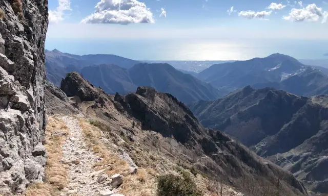 Sentiero roccioso su un crinale montuoso, con vista sul mare e sulle colline. Un percorso che ricorda la via Vandelli, un'antica strada panoramica.