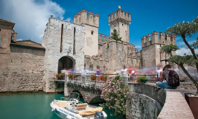 Castello Scaligero di Sirmione, un'ottima idea per cosa vedere a Sirmione in un giorno. Un ponte pedonale affollato porta all'ingresso del castello, con un motoscafo ormeggiato nell'acqua turchese.