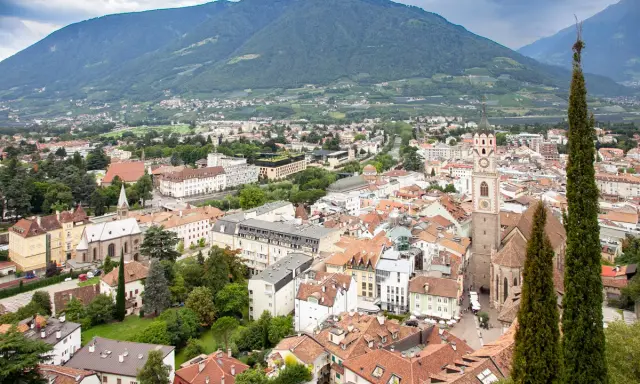 Vista aerea del centro storico di Merano, con montagne sullo sfondo e un campanile che svetta tra i tetti rossi.