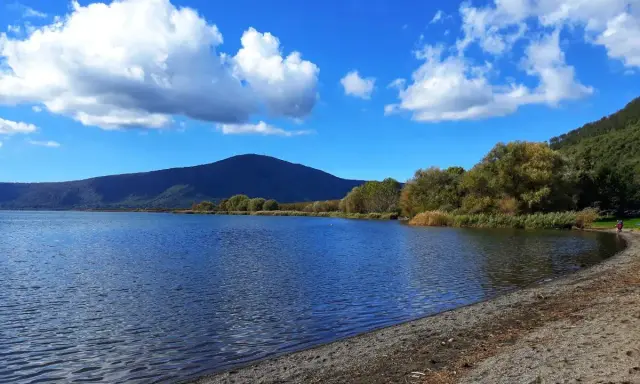 Spiaggia ghiaiosa e acque calme del lago di Vico, con montagne verdi e cielo azzurro punteggiato di nuvole bianche.