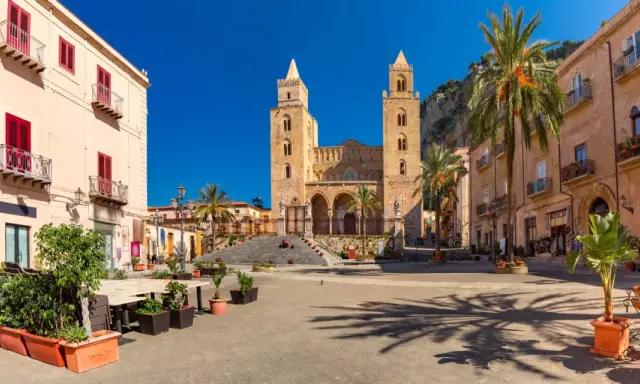 Piazza soleggiata con la cattedrale normanna sullo sfondo, palme e palazzi storici. Un luogo incantevole tra i borghi da visitare in Sicilia.