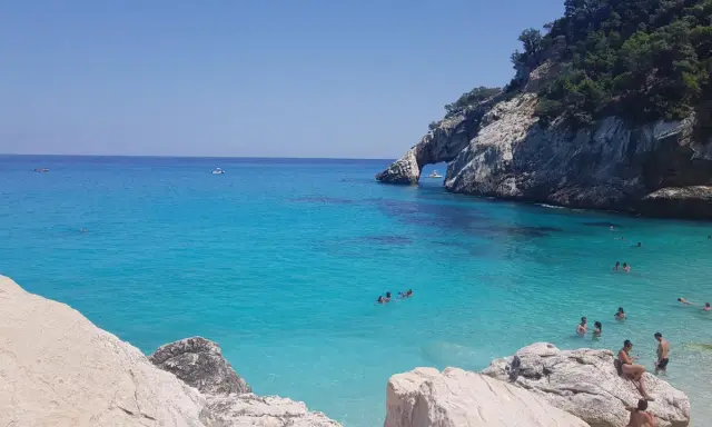 Acqua cristallina e scogliere a picco a Cala Goloritzè, raggiungibile con un suggestivo **percorso a piedi**. Persone nuotano e si rilassano.
