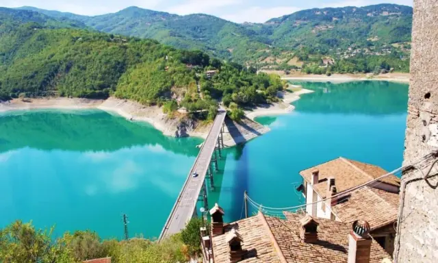 Panoramica mozzafiato del lago turchese vicino a Castel di Tora, con un ponte che attraversa le acque cristalline e montagne verdi sullo sfondo.