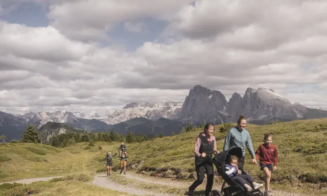 Famiglia in escursione sull'Alpe di Siusi, con bambini e passeggino, sullo sfondo le maestose vette dolomitiche.