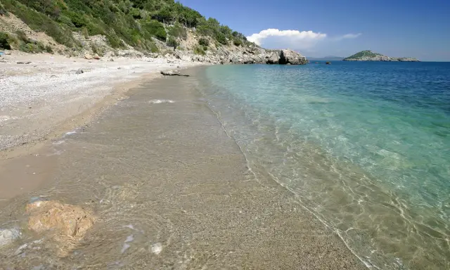 Spiaggia di ciottoli e sabbia con acqua cristallina, vista verso il mare aperto e un'isola in lontananza, vicino al porto di Ercole.
