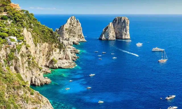 Scogliere verdi e rocciose si affacciano sul blu intenso del golfo di Napoli, punteggiato da barche e yacht. Faraglioni iconici emergono dal mare.