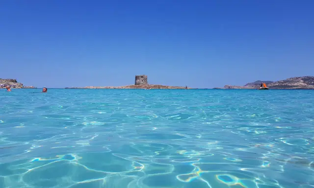 Acque cristalline del mare nord Italia con torre antica su isolotto e barca a vela arancione.