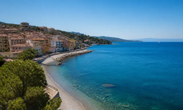 Porto Santo Stefano: case colorate affacciate su spiagge di ciottoli e sabbia, con il mare azzurro e un veliero all'orizzonte.