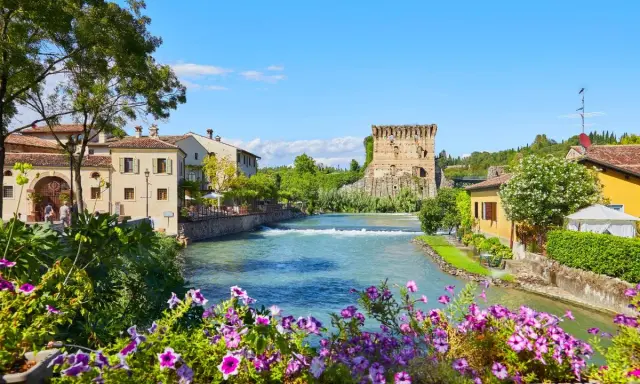 Un fiume attraversa un pittoresco borgo medievale veneto, con un'antica torre in rovina e case colorate.