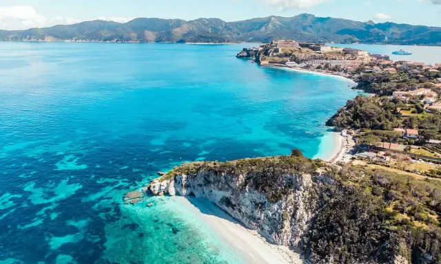 Costa rocciosa con spiaggia bianca e mare cristallino, un borgo fortificato domina la baia. Splendida località mare Toscana.