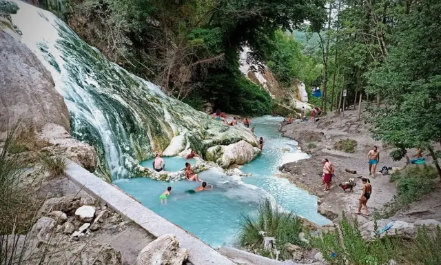 Cascata bianca e pozze azzurre alle terme San Filippo, con persone che si rilassano nell'acqua termale.