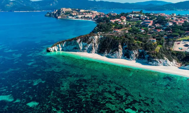 Vista aerea di una spiaggia bianca con acque cristalline e un borgo storico su una collina, parte dell'arcipelago toscano isole visitabili.
