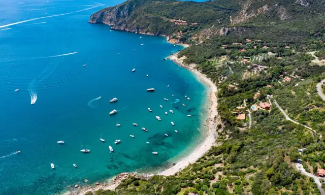Baia con acque cristalline e numerose barche, incastonata tra colline verdi e scogliere. Un vero gioiello tra le spiagge più belle Toscana.