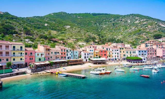 Colorate case affacciate sul mare cristallino, con barche ormeggiate e la collina verde sullo sfondo. Un'immagine che evoca il fascino del traghetto per l'Isola del Giglio.