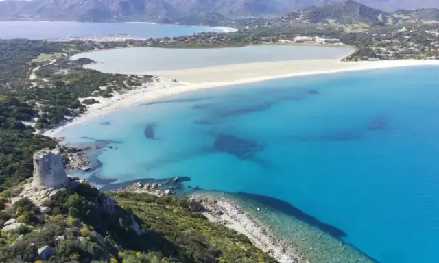 Torre costiera su promontorio verdeggiante domina una baia con acque cristalline, tra le spiagge più belle Sardegna.