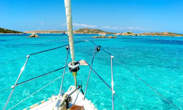 La prua di una barca a vela naviga verso La Maddalena, tra acque cristalline e isole rocciose.