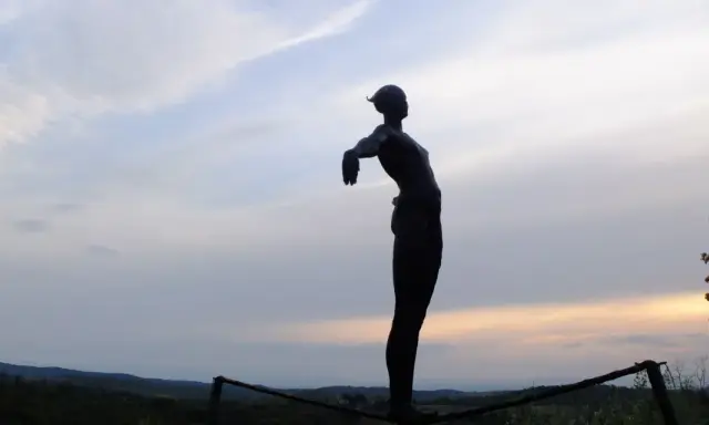 Statua silhouette con braccia aperte, su un crinale collinare, al tramonto. Un punto panoramico lungo le tappe della Via Romea Sanese.