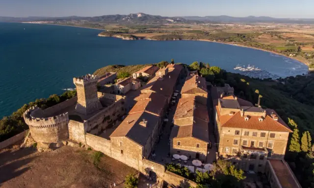 Borgo medievale fortificato domina la campagna toscana lungo il mare, con un castello imponente e case dai tetti rossi affacciate su un golfo con barche a vela.