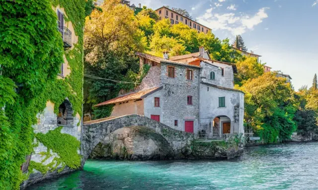 Antichi borghi medievali lombardia da visitare: un ponte di pietra collega case storiche sull'acqua cristallina, con edera rampicante e vegetazione lussureggiante.