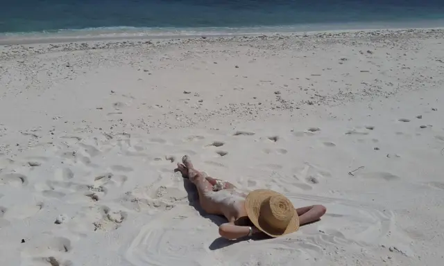 Persona distesa su sabbia bianca, con cappello di paglia, vicino al mare turchese. Un'immagine che evoca la pace delle spiagge naturiste in Italia.