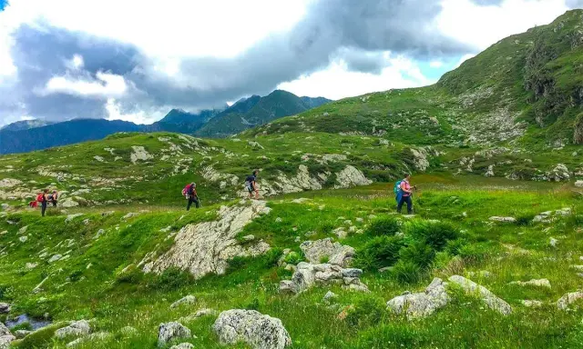 Escursionisti attraversano un paesaggio verdeggiante in una località montagna Piemonte, con rocce e nuvole minacciose all'orizzonte.