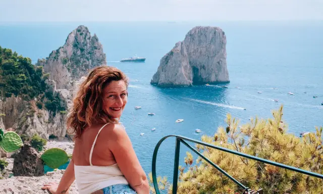 Donna sorridente con vista sui Faraglioni, un'ispirazione per come visitare Capri. Mare azzurro e barche in lontananza.