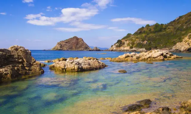 Acque cristalline e rocce scolpite dal mare a Porto Ercole, con montagne verdi sullo sfondo e un cielo azzurro punteggiato di nuvole.
