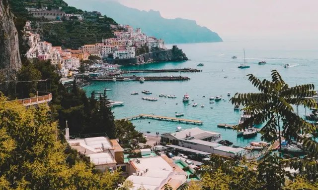 Vista panoramica di un villaggio colorato sulla Costiera Amalfitana, con barche nel porto e montagne sullo sfondo. Un invito a scoprire come visitare la Costiera Amalfitana.