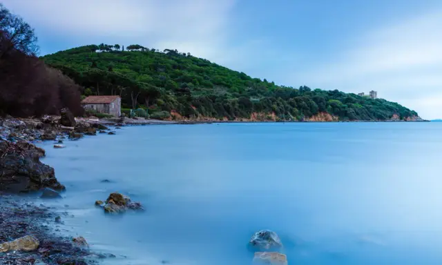 Una casa di pietra sulla spiaggia di Bengodi, con il mare calmo e una collina verdeggiante sullo sfondo.