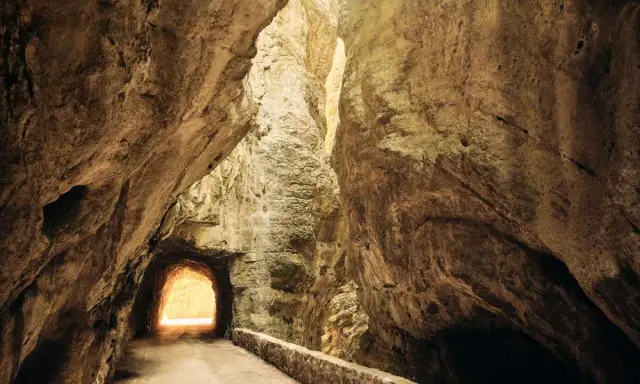 La strada della forra si snoda tra imponenti pareti rocciose, con un tunnel che si apre verso una luce accecante.
