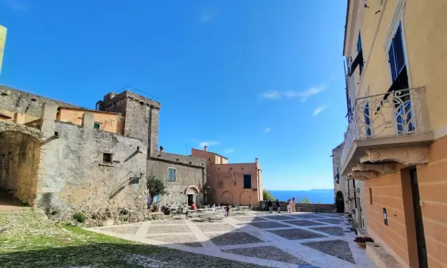 Piazza nel centro storico di Borgio Verezzi con edifici antichi, tavolini all'aperto e il mare sullo sfondo.